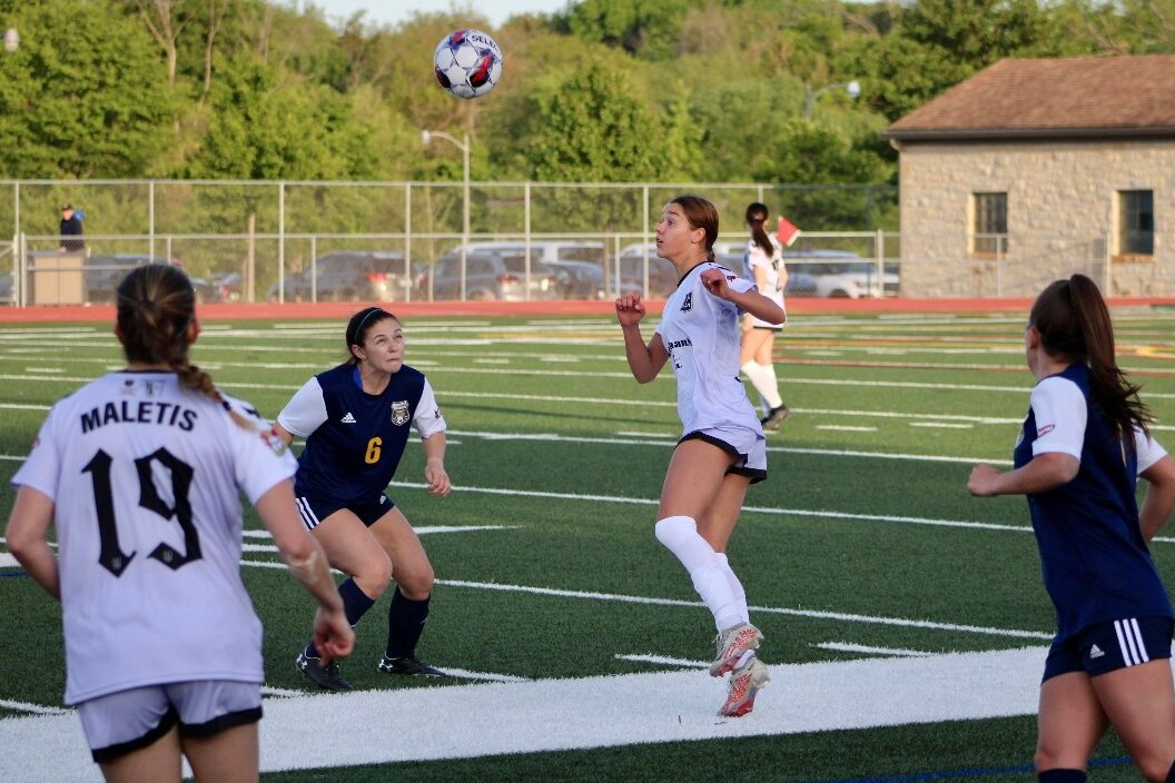 Left of frame, Alexa Maletis 19, with her back to the camera. Center frame: two players contest an aerial ball (one in navy kit, one in all white kit)