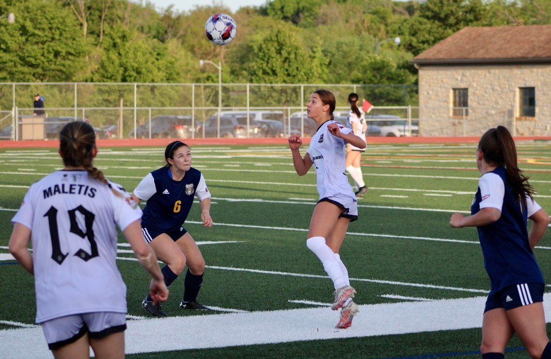Left of frame, Alexa Maletis 19, with her back to the camera. Center frame: two players contest an aerial ball (one in navy kit, one in all white kit)