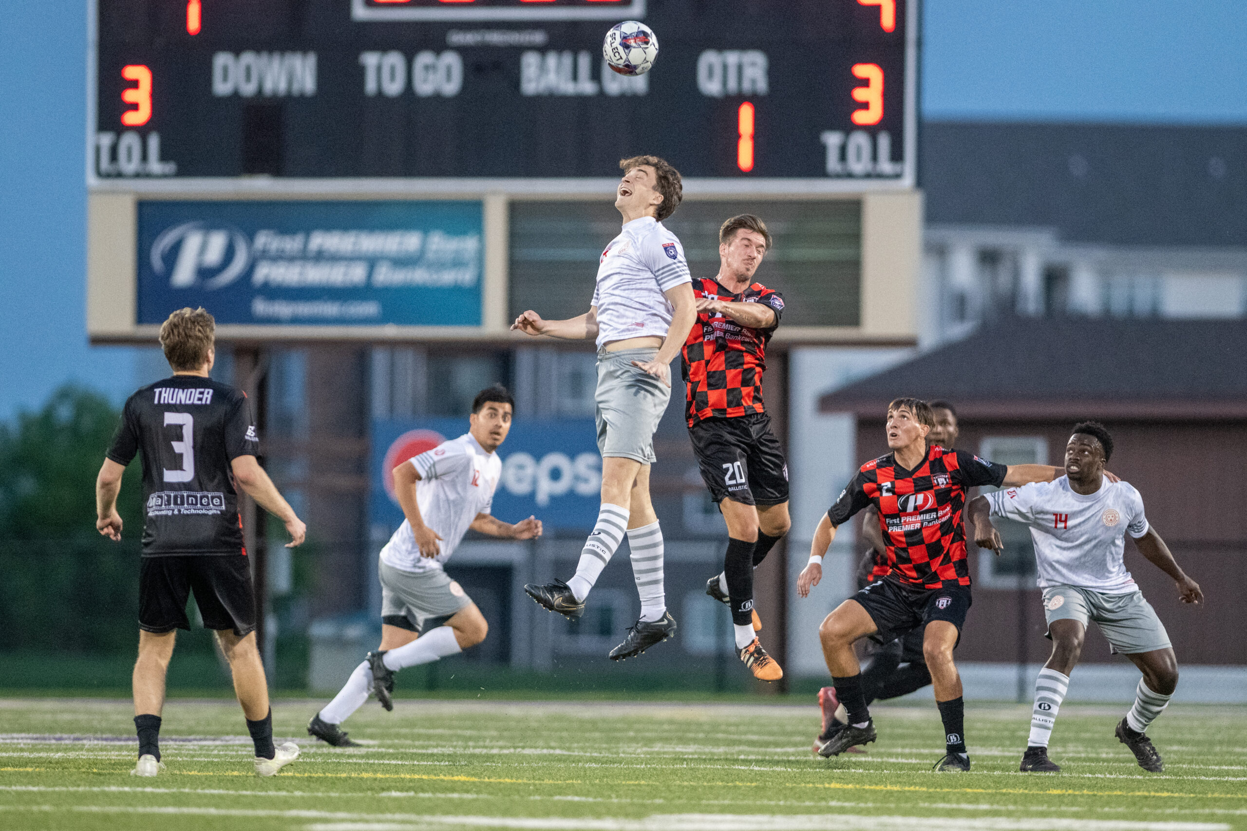 Players in all white and in red and black checked soccer kits are pictured in mid-air