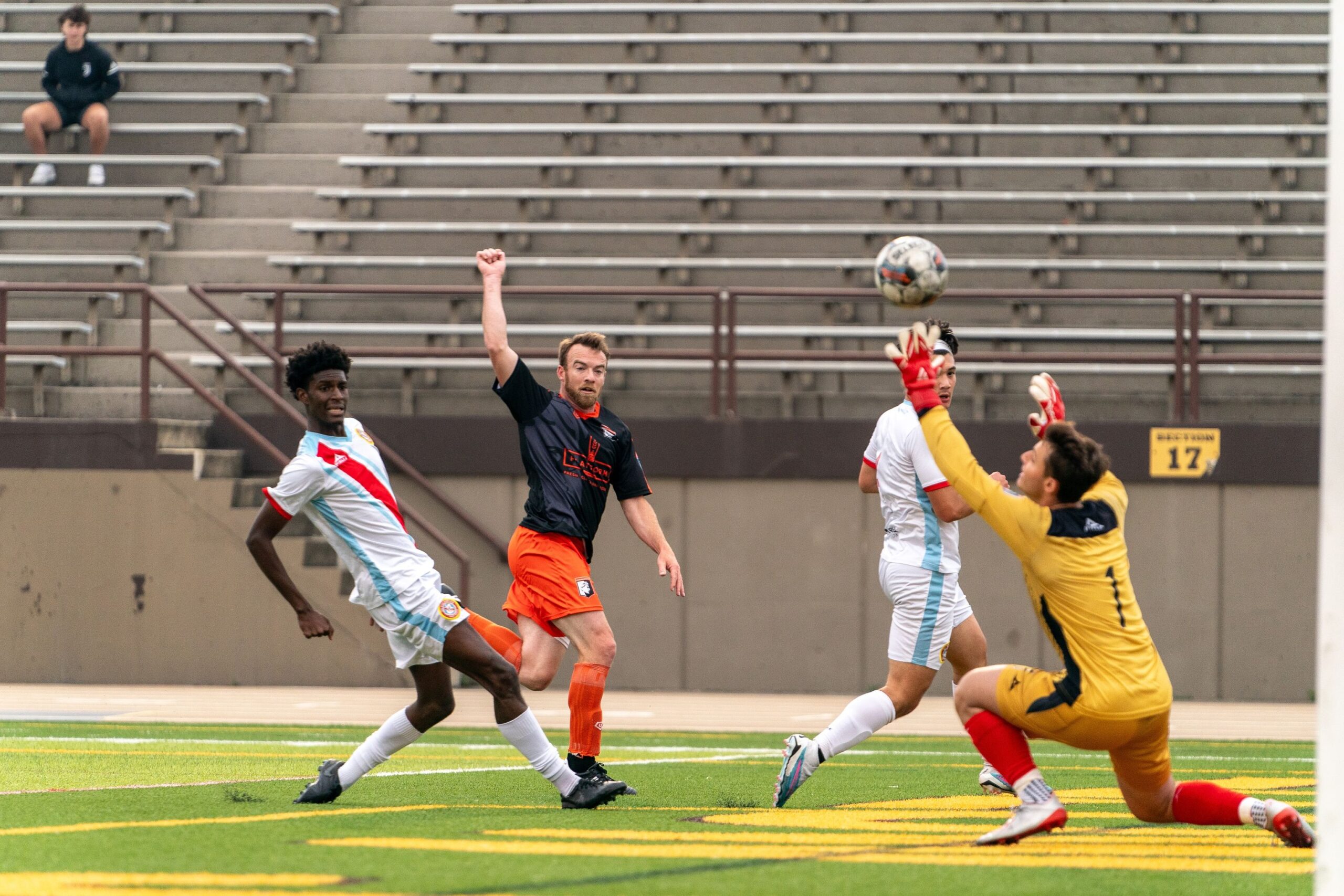 Four soccer players are depicted. Left hand side, player in a white jersey sliding in, player in center in a black jersey and orange shorts with his arm aloft, partially obscured player in white jersey and a goalkeeper on the right side, diving towards the ball