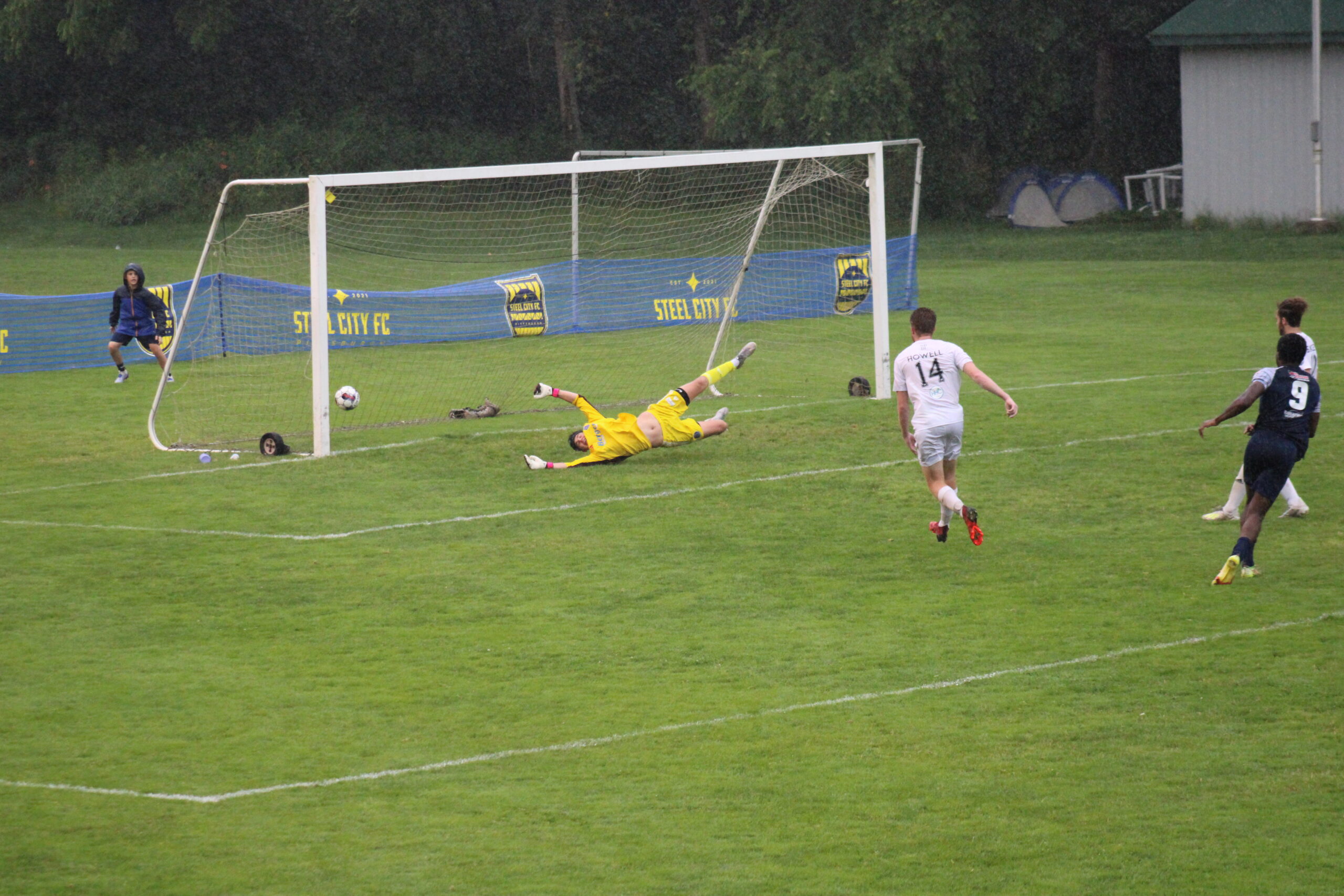 The ball is in the center-left of the frame, just inside the goal. Yellow kitted goalkeeper is horizontal across the goal. White kitted defender is in center of frame. Black kitted attacker is in right of field