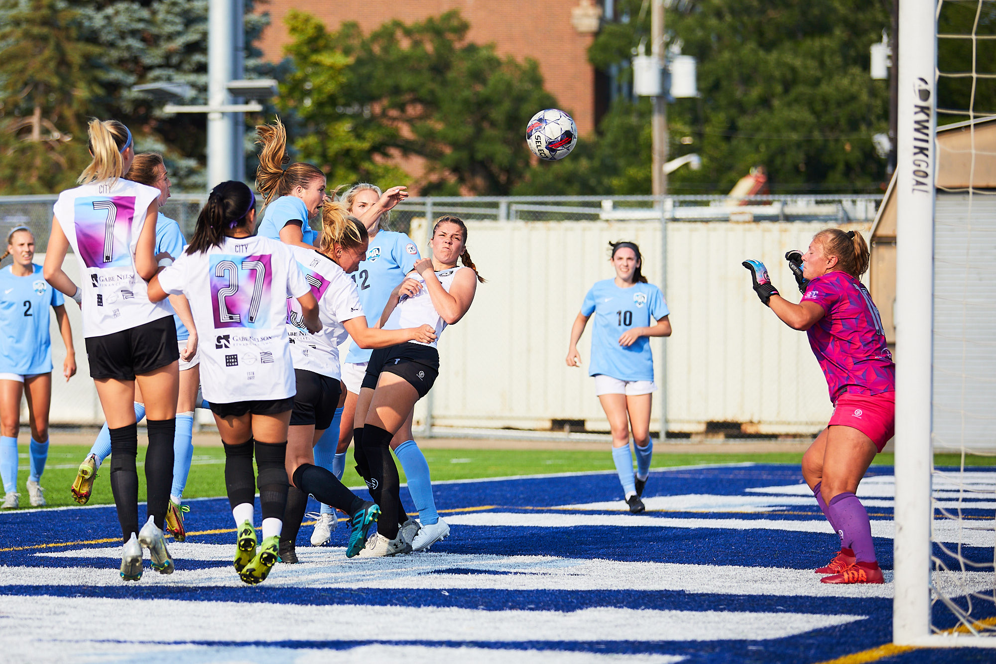 A group of women in soccer kits fight for a ball, which is in the middle of the frame. A goalkeeper is pictured in the right side, diving